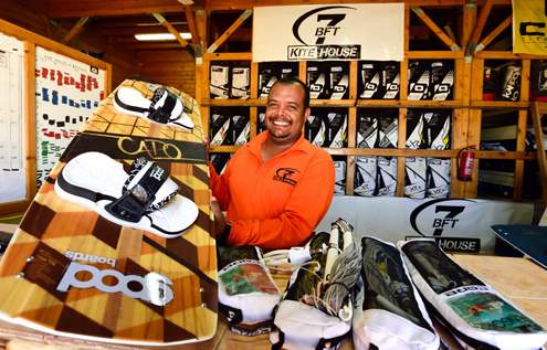 A smiling staff member at the KiteHouse in Somabay, holding a kiteboard, surrounded by neatly organized kite equipment inside a wooden rental shop.