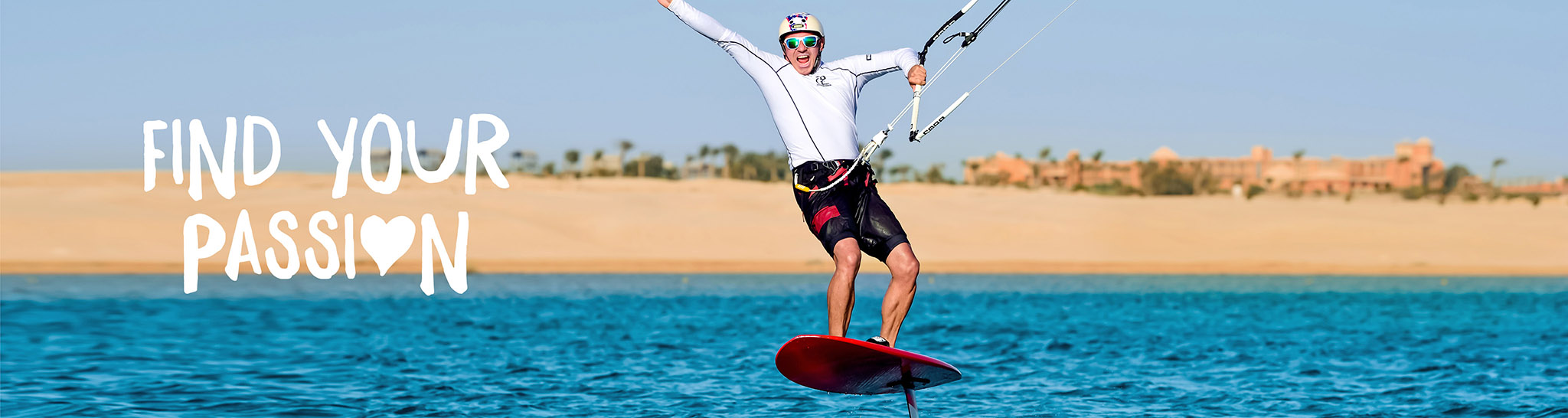Kitesurfer hydrofoiling over the blue sea, arms raised in excitement, with a desert landscape and resort in the background.