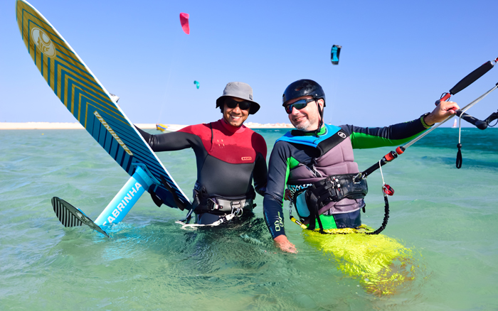 Zwei Kitesurfer stehen im seichten Wasser, lächeln und halten ihre Bretter, während im Hintergrund in der Soma-Bucht bunte Drachen fliegen.