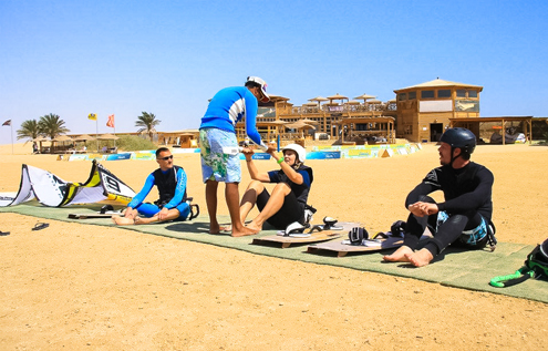 A kitesurfing instructor teaching three students on a mat at Kitehouse Somabay, with the wooden beachfront center in the background.