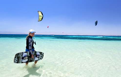 Eine Kitesurferin steht mit ihrem Board im seichten, kristallklaren Wasser und blickt lächelnd in die Kamera. Im Hintergrund gleiten mehrere Kitesurfer über das türkisfarbene Meer unter strahlend blauem Himmel.