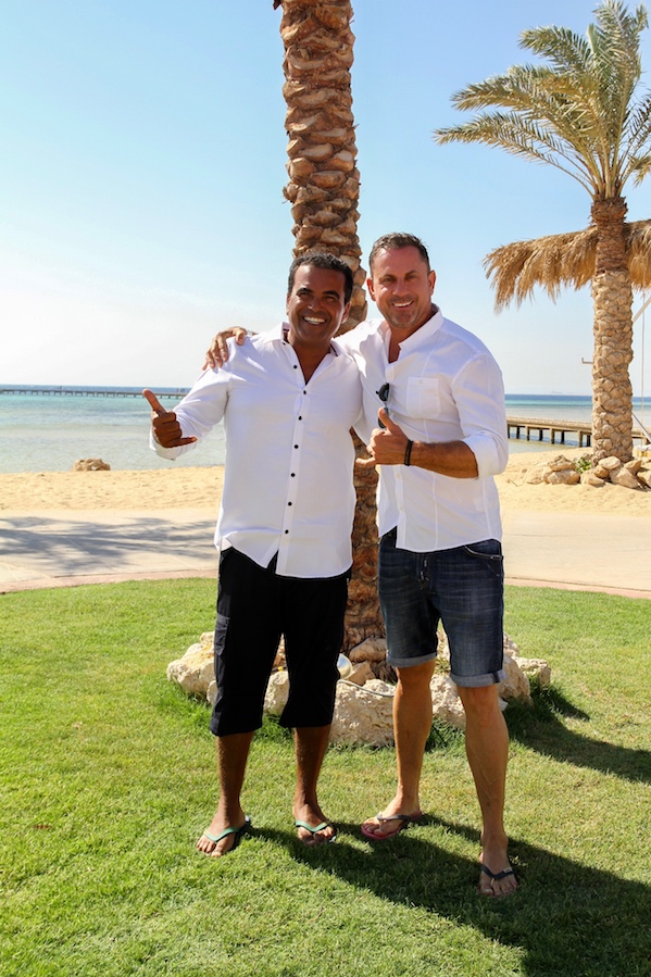 Two men dressed in white shirts and casual summer clothes are standing on a green lawn near the beach, smiling and giving the thumbs up. In the background are palm trees, a turquoise sea and a wooden pier.