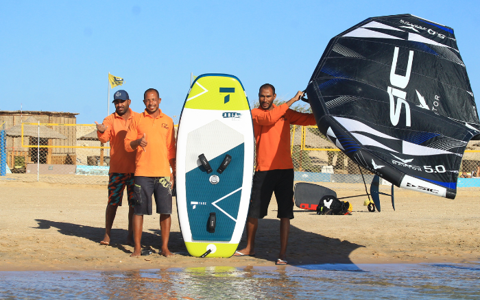 Drei Ausbilder in orangefarbenen Hemden stehen am Strand von Soma Bay, in der Hand ein Wingfoil-Brett und einen schwarzen SIC-Flügel.