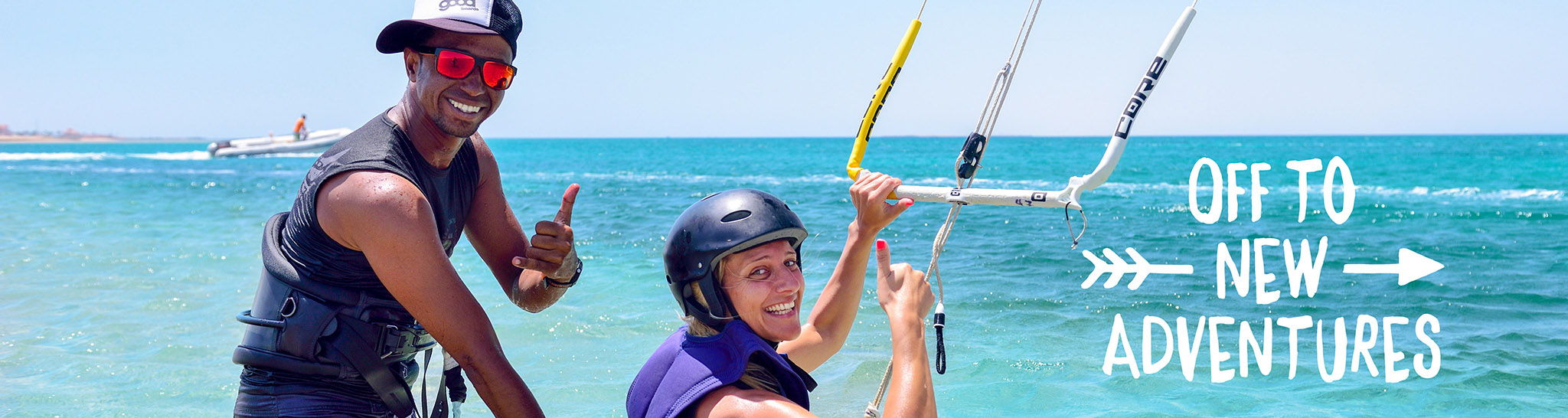 Kitesurf instructor and student smiling and giving thumbs-up in shallow turquoise water. The student holds a kite bar, ready for a new adventure. The background features the open sea, a clear blue sky, and a rescue boat.