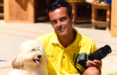 Ein Mann im gelben Polo-Shirt mit Sonnenbrille auf dem Kopf hält eine Kamera in der Hand und sitzt neben einem weißen, flauschigen Hund. Beide genießen die Sonne vor einer Holzkulisse im KiteHouse.
