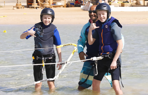 Two young kitesurfing students in wetsuits and helmets learning kite control in shallow water with their instructor at Kitehouse Somabay.