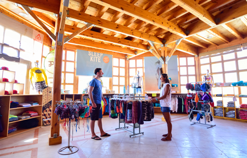 Ein Mann und eine Frau stehen im KiteHouse Surf Shop, umgeben von Kitesurf-Bekleidung, Accessoires und Ausrüstung. Der helle Raum mit Holzkonstruktion und großen Fenstern sorgt für eine entspannte Shopping-Atmosphäre.