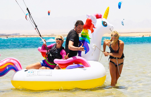 Three people enjoying the water at KiteHouse Somabay with a giant inflatable unicorn, while kitesurfers glide over the sea in the background.
