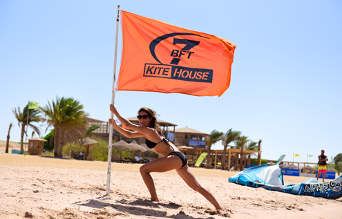 Eine Frau im Bikini hält eine orangefarbene 7 Bft KiteHouse-Flagge am Strand von Somabay, während im Hintergrund Kites und Palmen zu sehen sind.
