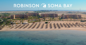 Aerial view of Robinson Soma Bay, a beachfront resort in Egypt, featuring a sandy beach with sun loungers, clear blue sea, and resort buildings in the background.